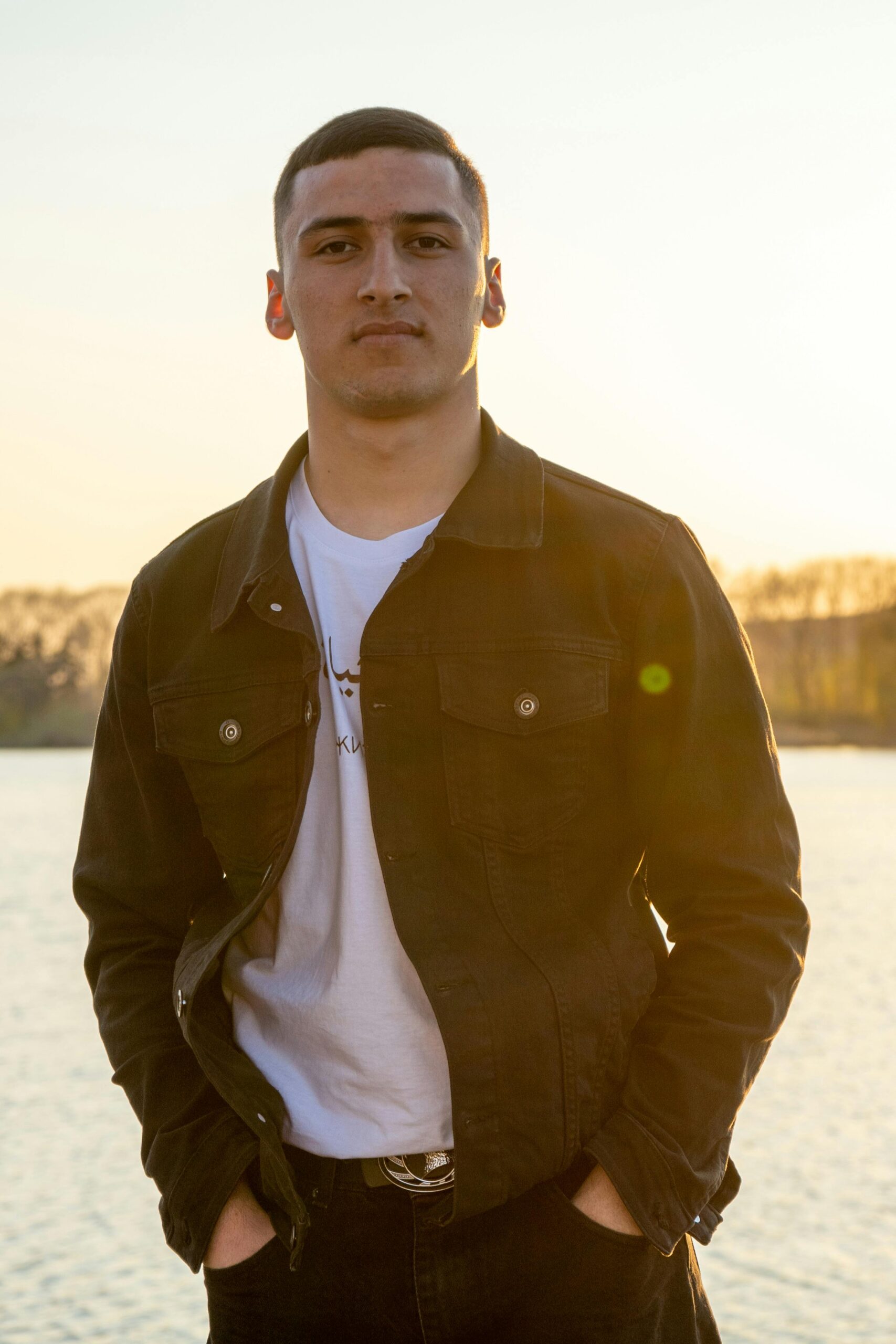 Young man in casual attire stands confidently by the water during sunset.