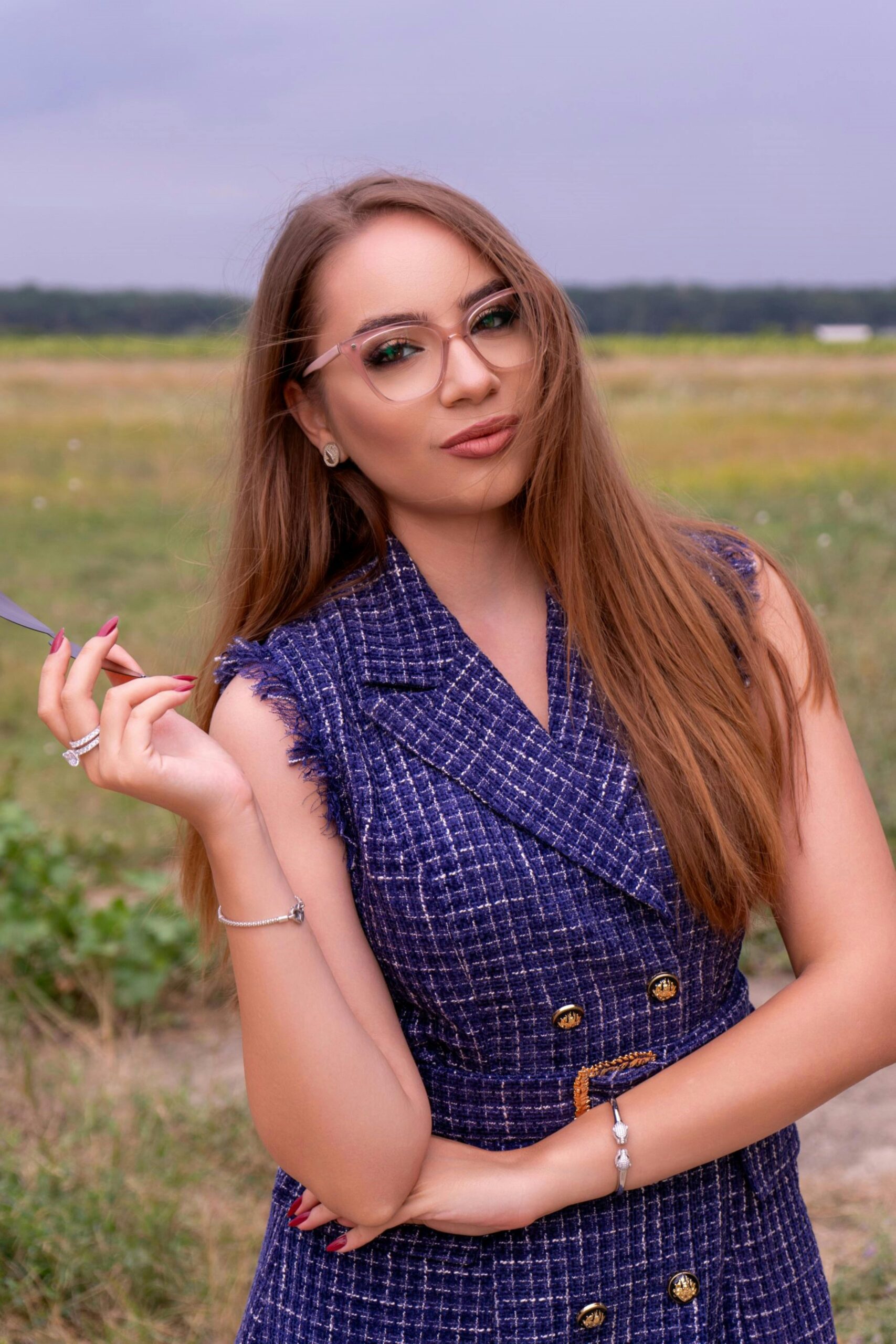 A fashionable young woman with glasses posing outdoors in a field setting during summer.